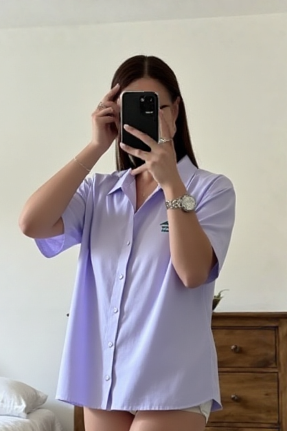 Person wearing a light purple shirt taking a mirror selfie in a bedroom.